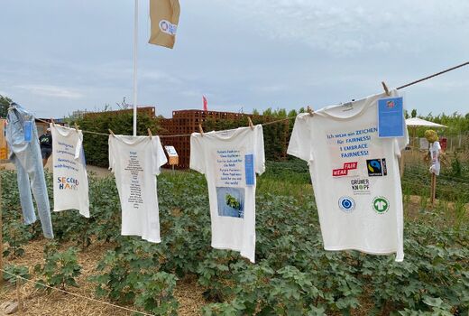 Man sieht T-Shirts auf einer Wäscheleine über einem Feld. Auf den T-Shirts sind Fair Trade Logos zu sehen.