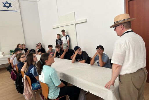 Die SchülerInnen sitzen an einem langen Tisch. Vorne steht ein Mann und spricht.