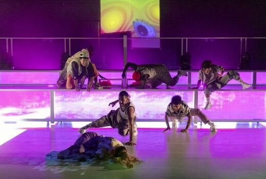 Auf einer stufigen, lilafarbenen Tribüne bewegen sich fünf Schauspieler auf dem Boden. Links im Vordergrund liegt eine gekrümmte Gestalt auf dem Boden.