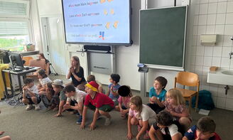 Eine Gruppe von Kindern hockt auf dem Boden. Im Hintergrund sieht man einen kurzen Liedtext auf einer digitalen Tafel. Die Lehrerin sitzt hinter den Kindern und hat den Zeigefinger an den Lippen als Ruhesymbol.