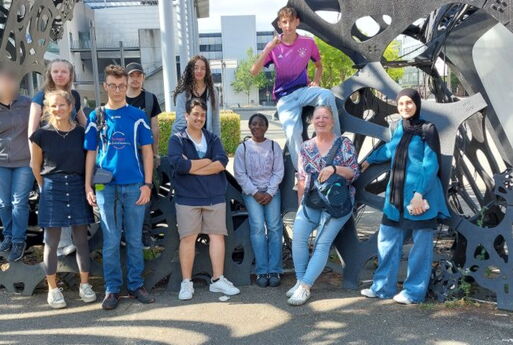 Neun Schüler und zwei Lehrerinnen posieren vor einem großen Kunstwerk aus Metall-