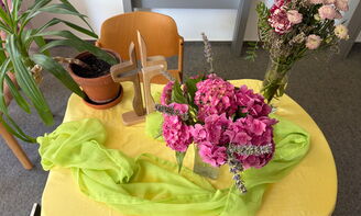 Auf einem Tisch liegt eine gelbe Decke, darauf stehen mittig und recht Blumenvasen mit pinken und rosa Blumen drin. Hinten links steht eine Zimmerpflanze und rechts daneben ein Kreuz aus Holz. Außerdem liegt dort ein gelb-grünes Tuch.