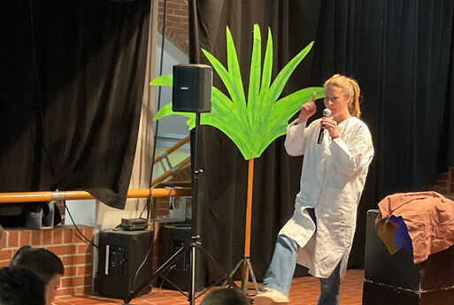 Eine Lehrerin steht auf einer Bühne und hebt ihr rechtes Bein und den rechten Finger nach oben, links neben ihr im Bild sieht man Musikboxen und eine Palme aus Pappe