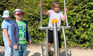Ein Kind nutzt ein Fitnessgerät und schaut in die Kamera. Zwei weitere Kinder stehen an.