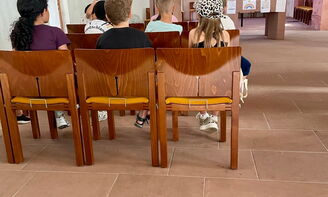 Kinder sitzen auf Stühlen in einer Kirche und schauen nach vorne. Dort sprechen zwei Frauen in Mikrofone. Hinter den Kindern liegt auf dem Boden ein Schulhund.