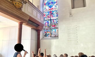 Eine große Kindergruppe steht unterhalb einer Orgel und betrachtet mit einer Pfarrerin ein Kirchenfenster.