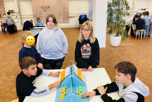 Zwei Jungs sitzen sich an einem Tisch gegenüber und lösen Aufgaben zur Geheimschrift. Drei Mädchen stehen am Tisch und schauen ihnen zu.