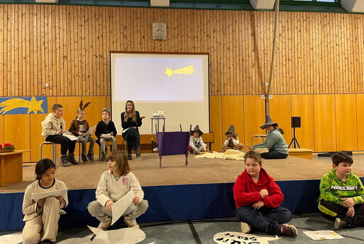 Zehn Kinder der dritten Klasse führen die Weihnachtsgeschichte erzählt vom Esel Jonas auf. Vier Kinder sitzen vor der Bühne. Drei Kinder links auf der Bühne auf Stühlen und drei Kinder rechts auf dem Boden. In der Mitte sieht man eine Krippe.