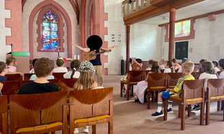Eine große Kindergruppe sitzt auf Stühlen in einer Kirche. Vor der Gruppe steht eine Pfarrerin mit Gitarre.