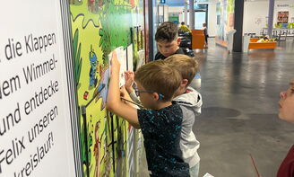 Vier Kinder stehen mit Büchern und Stiften in der Hand an oder vor einer bunten Wand mit Text, manche schreiben in ihr Buch
