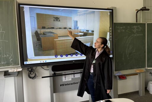 Eine junge Frau im Talar steht vor einem interaktiven Board mit dem Bild eines Gerichtssaals und zeigt auf die Richterbank