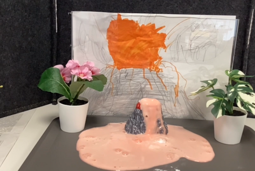In der Mitte des Bildes sieht man einen Mini-Vulkan auf einem Tablett stehen, aus dem Vulkan kommt eine schaumige Flüssigkeit raus, im Hintergrund sieht man ein selbst gemaltes Bild von einem Vulkan, links und rechts stehen Blumen