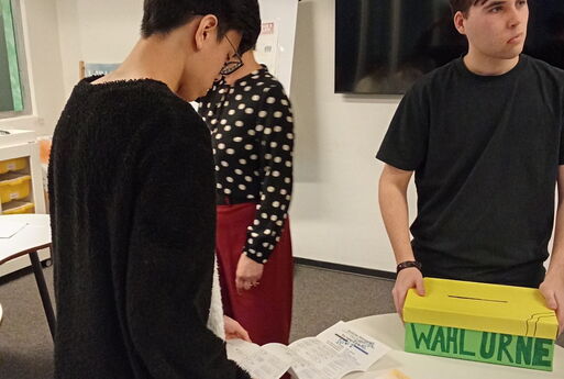 Ein Schüler schaut sich den Stimmzettel an. Ein anderer hält die Wahlurne fest.