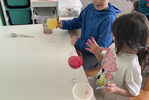 Zwei Kinder sitzen an einem Tisch, vor ihnen stehen zwei Gläser mit einer bunten Flüssigkeit drin, die Kinder schauen auf die Finger von dem Kind, das weiter vorne im Bild sitzt, die Finger sind rot gefärbt, im Hintergrund ist ein Klassenzimmer zu sehen