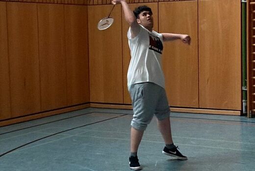 Ein Spieler schaut in die Luft und macht eine Schlagbewegung mit dem Badminton Schläger. Im Hintergrund ist die braune Wand der Turnhalle zu sehen.